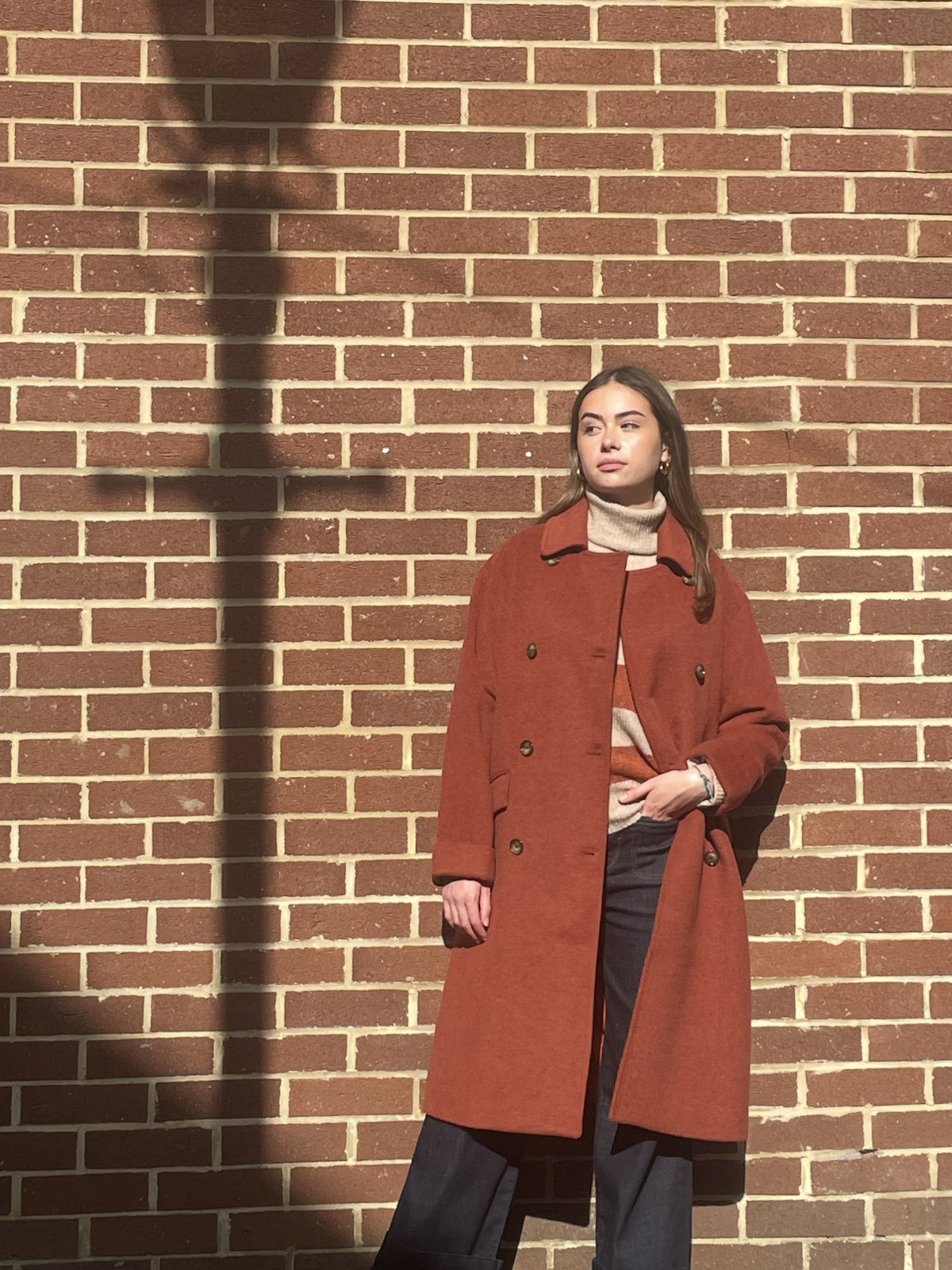 Model in a long rust double-breasted coat over a striped knit against a brick wall