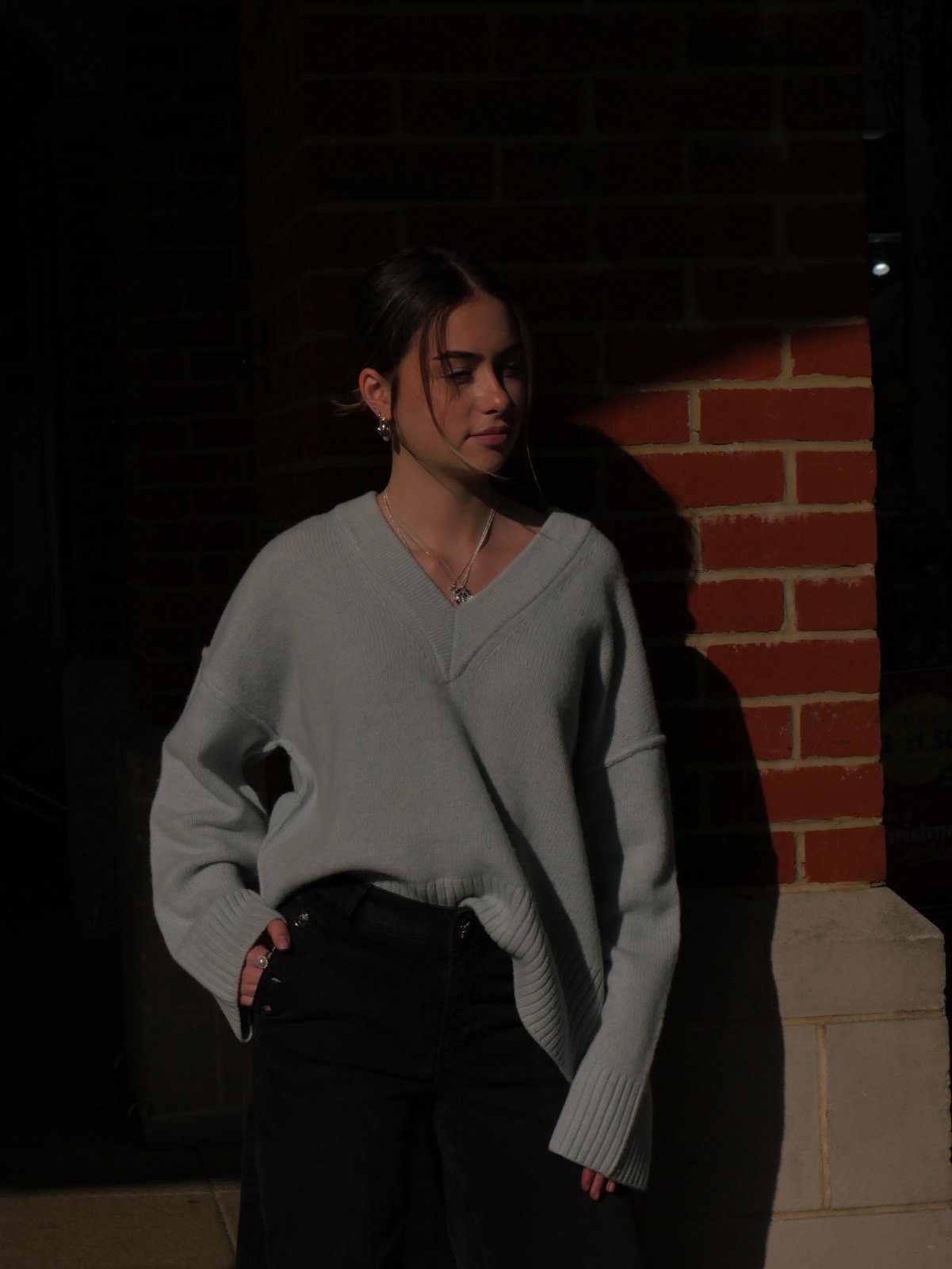 Model in a pale blue-grey V-neck jumper and black trousers, lit by a strip of sun against brick