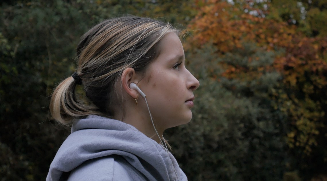 Profile portrait of Isabel Logan in a grey hoodie and earphones against autumn trees