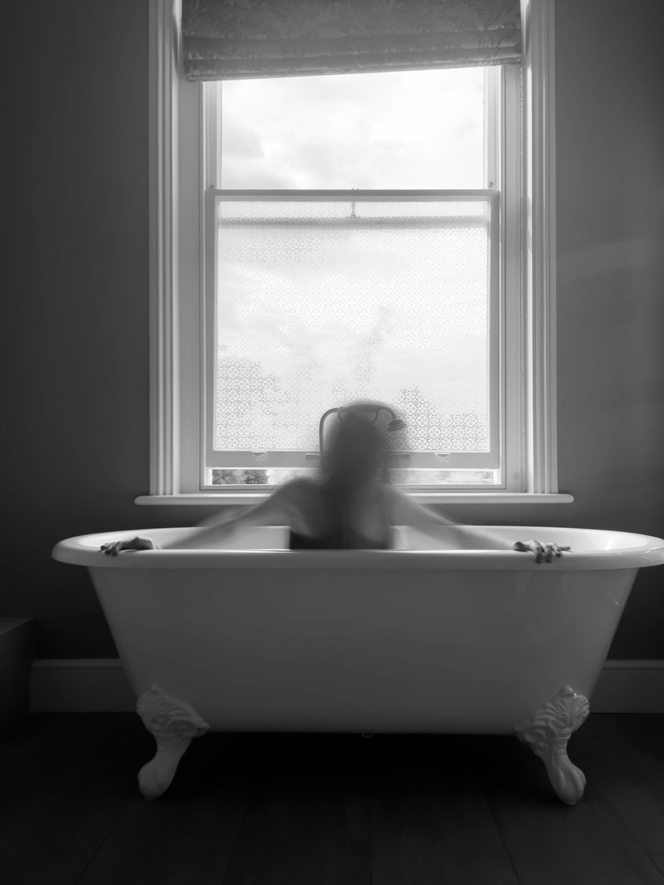 Black and white long-exposure of a blurred figure in a clawfoot bath in front of a sash window