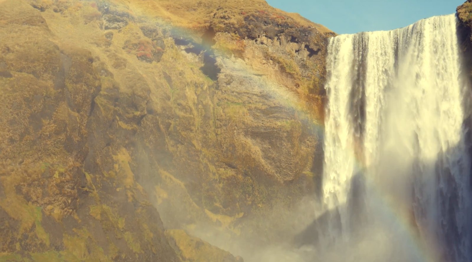 Still from Iceland 2023: a waterfall and rainbow beside a mossy cliff