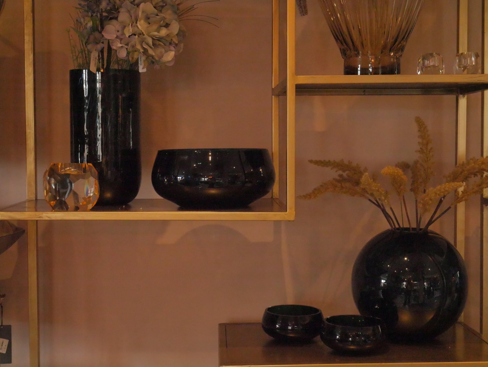 Black glass vases and bowls on gold shelving against a dusky pink wall