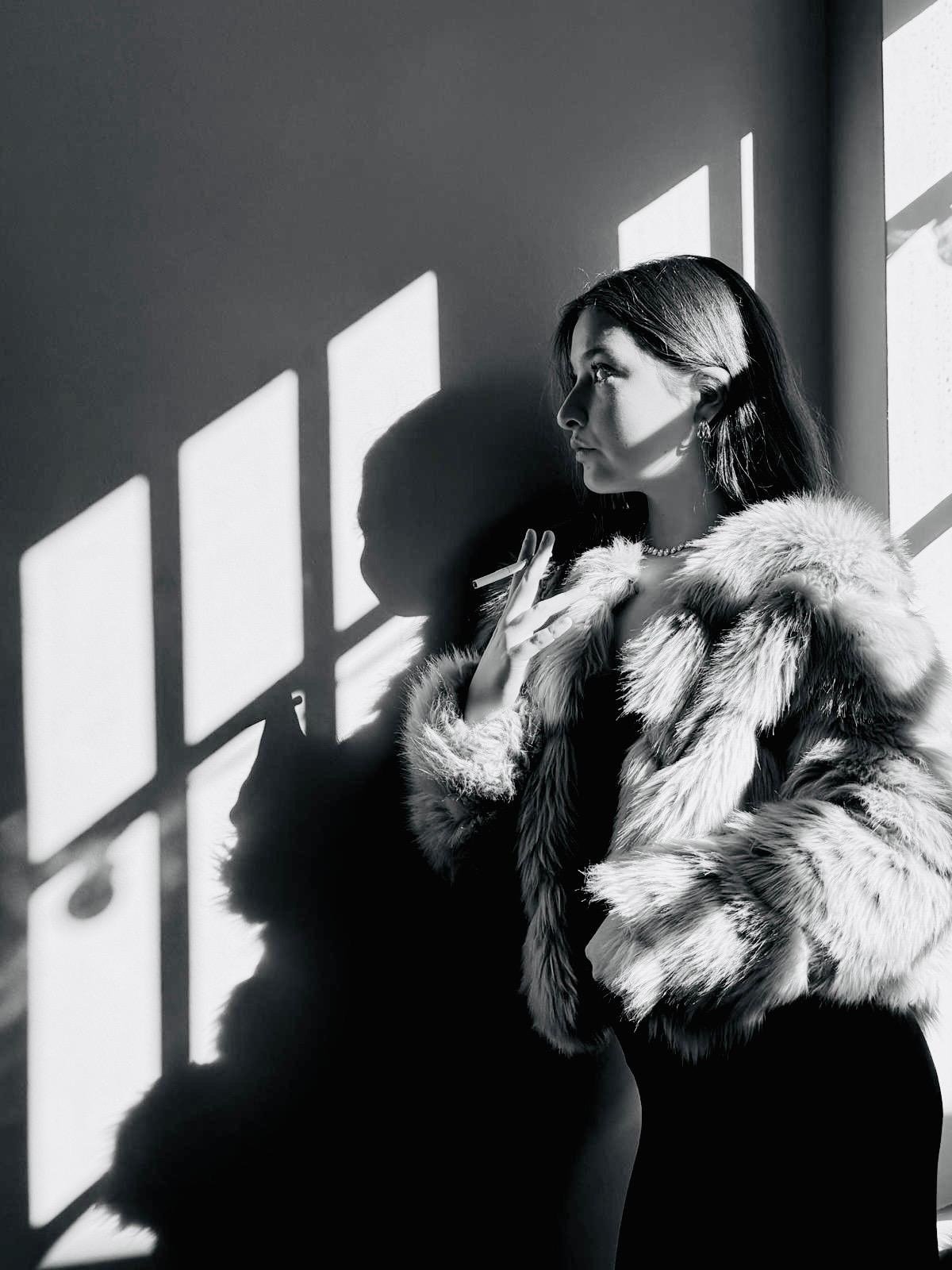 Black and white portrait of a woman in a fur coat holding a cigarette, lit by window shadows on the wall